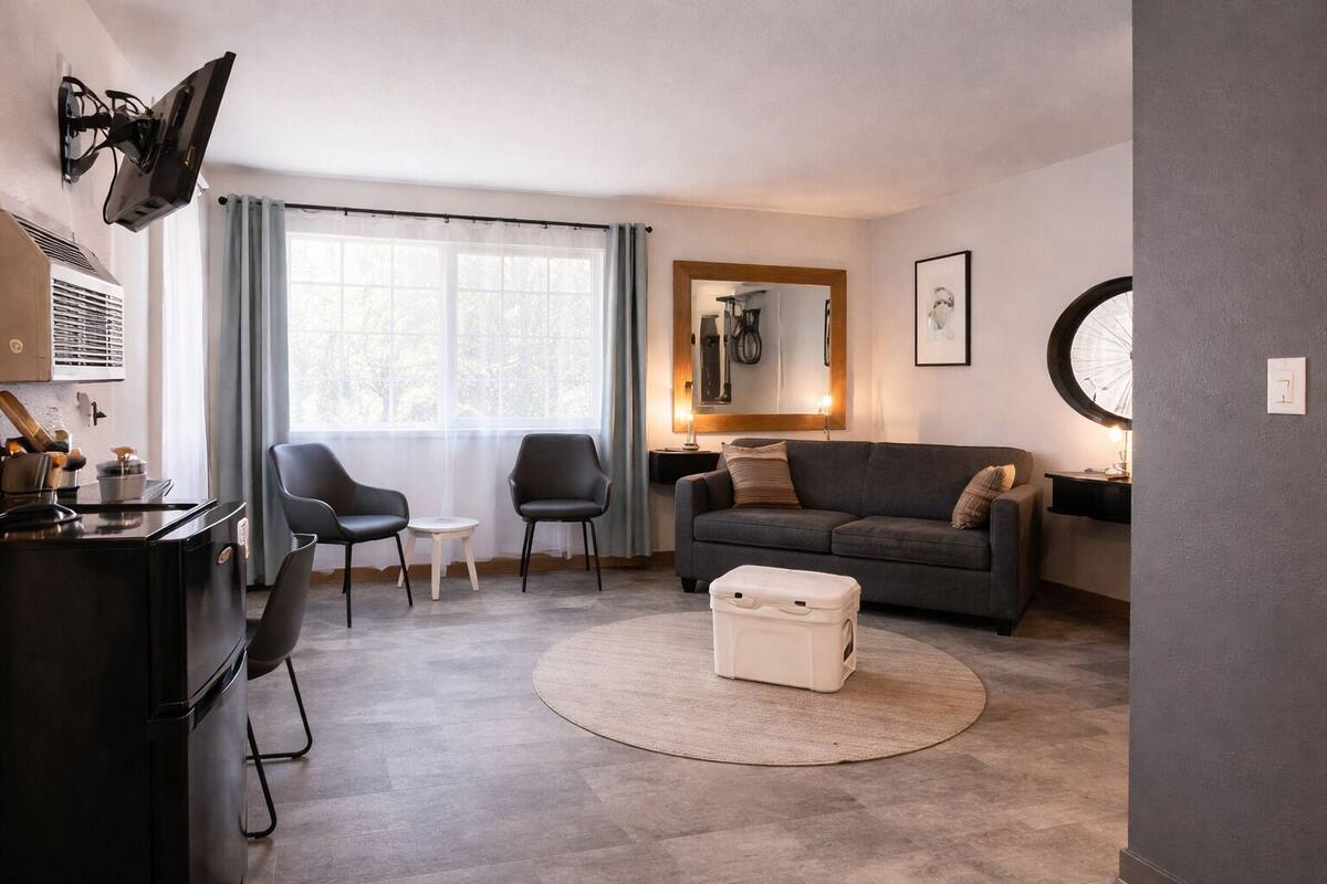 A modern living room with a sofa, two chairs, a small table, a TV mounted on the wall, and a large window with curtains.