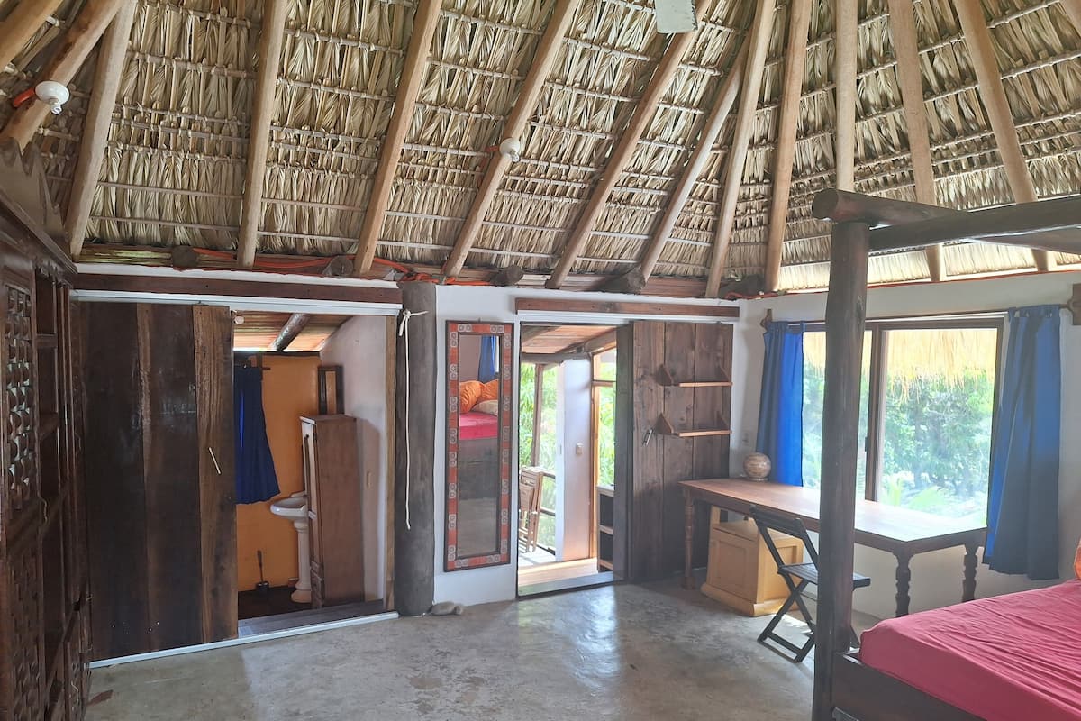 A rustic room with a thatched roof, wooden furniture, and a bed with a red bedspread.