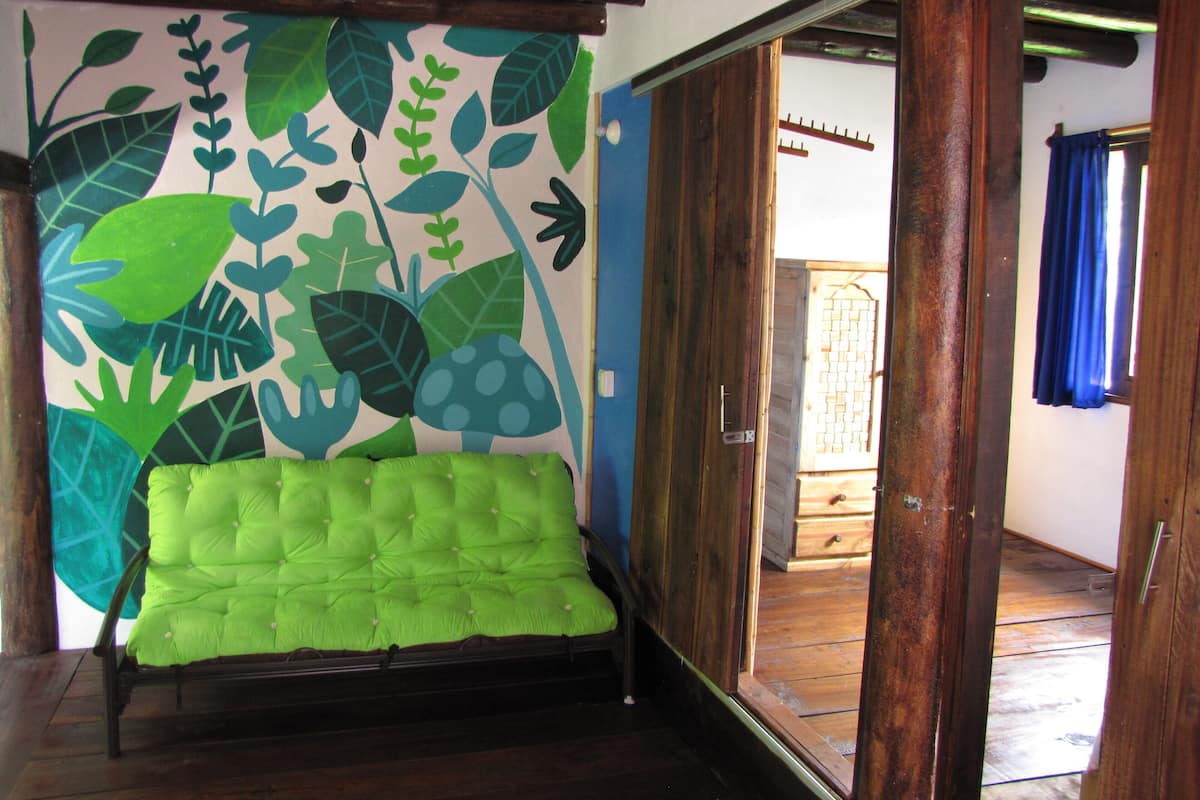 A room with a green tufted sofa, a wooden floor, a wall with a leaf pattern, and a wooden door leading to another room.