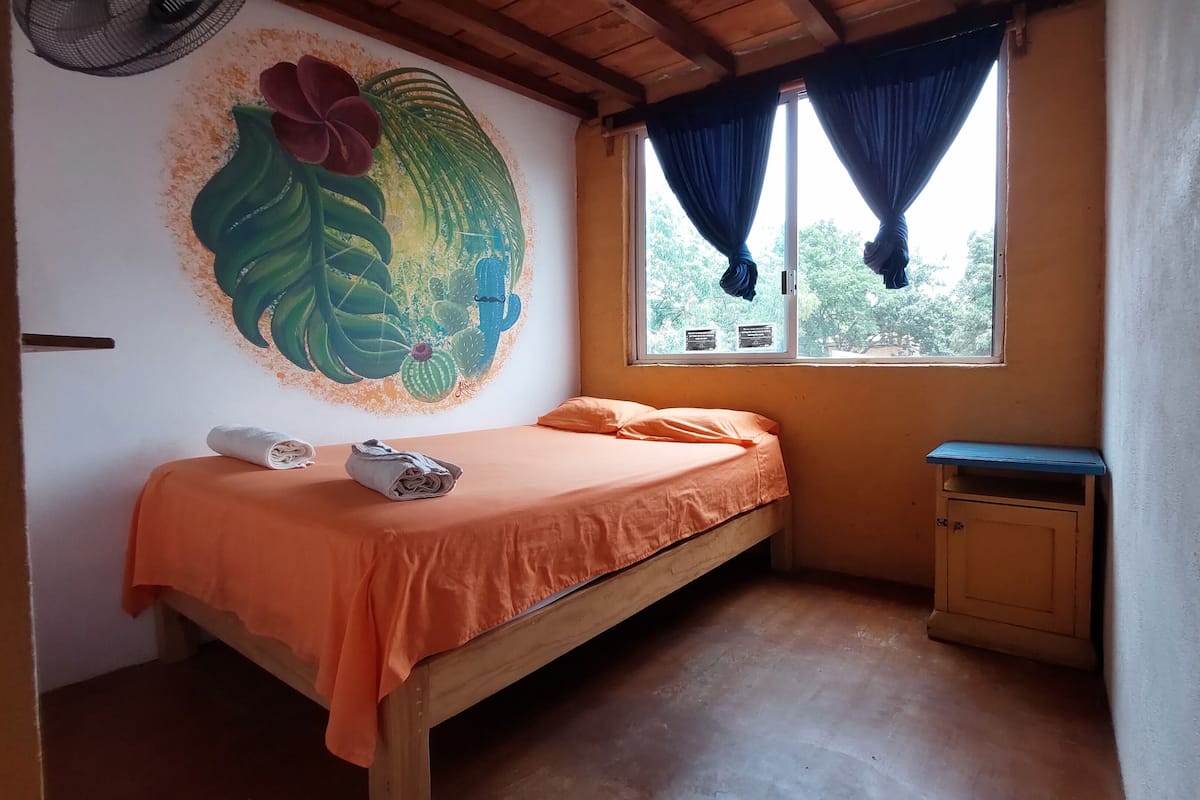 A small room with a single bed, a wooden cabinet, a window with curtains, and a wall mural.