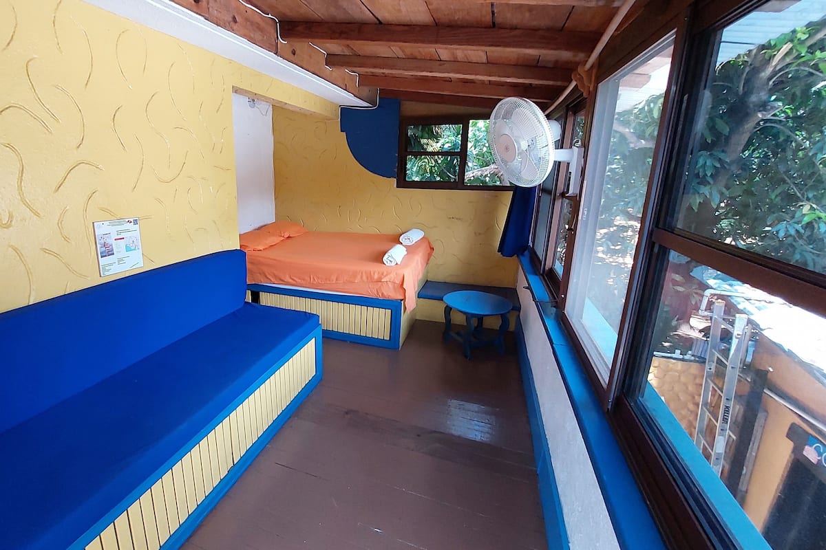A room with a bed, a blue bench, a small table, and a window with a view of greenery.