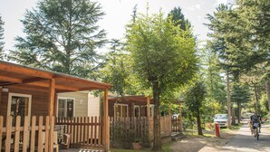 A row of wooden cabins with a wooden fence, a paved path, and a cyclist on the road.