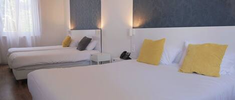 Une chambre d’hôtel avec trois lits simples, chacun doté de draps blancs et d’oreillers jaunes. On y trouve une petite table de chevet sur laquelle se trouve un téléphone. La chambre présente un design moderne, avec un grand mur d’accent de couleur grise et une fenêtre ornée de rideaux légers.