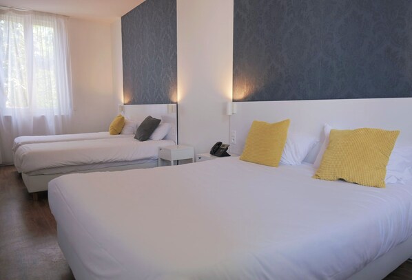 A hotel room with three single beds, each with white bedding and yellow pillows. There is a small bedside table with a phone on it. The room has a modern design with a large gray accent wall and a window with sheer curtains. - Green Hôtels Bordeaux Arena (Latresne)