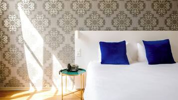 Una habitación de hotel con papel tapiz estampado, una cama blanca con almohadas azules, una mesita de noche verde pequeña con un teléfono y suelo de madera.
