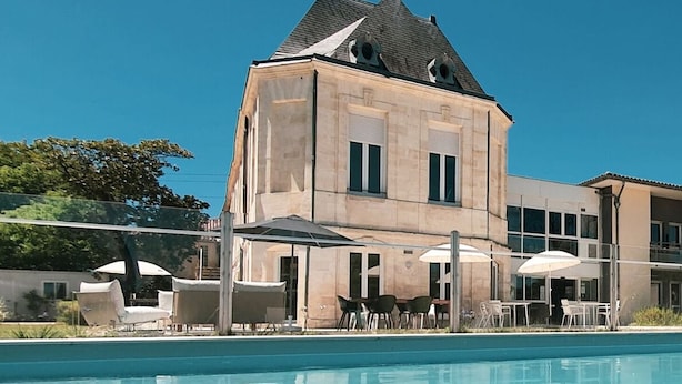 Ein Pool mit Liegestühlen und Sonnenschirmen, ein Steinhaus mit steilem Dach und ein klarer, blauer Himmel.
