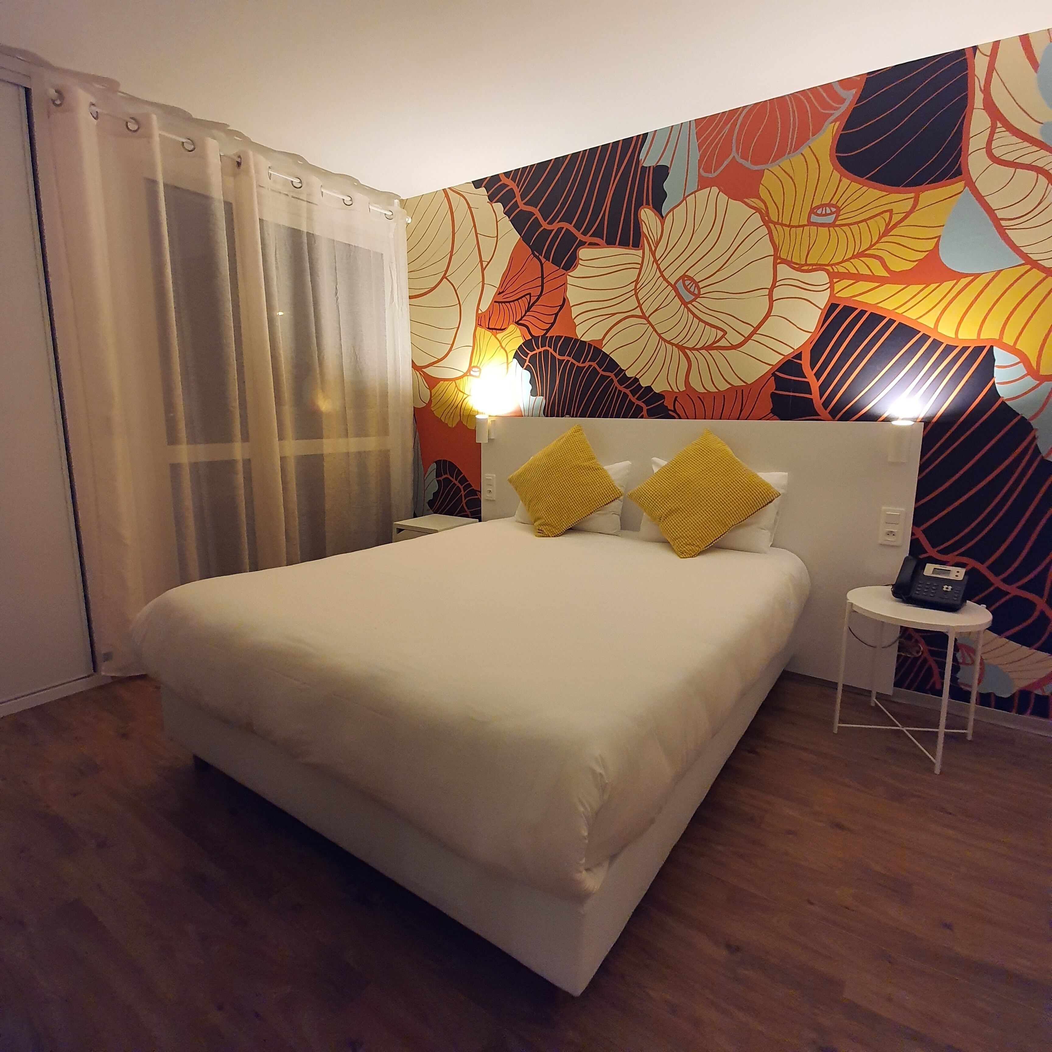 A hotel room with a large bed, a bedside table with a phone, and a colorful floral wall mural.