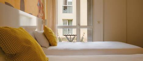 Quarto de hotel com cama, dois travesseiros amarelos, janela e vista para um edifício.