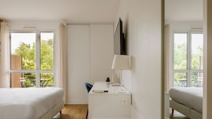 Ein Hotelzimmer mit zwei Betten, einem Schreibtisch mit Lampe und Blick auf Grünanlagen durch die Fenster.