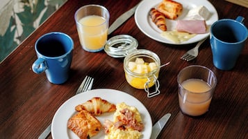 A wooden table with breakfast items including croissants, scrambled eggs, ham, and toast. There are two blue cups, a glass of orange juice, and a glass of tea.