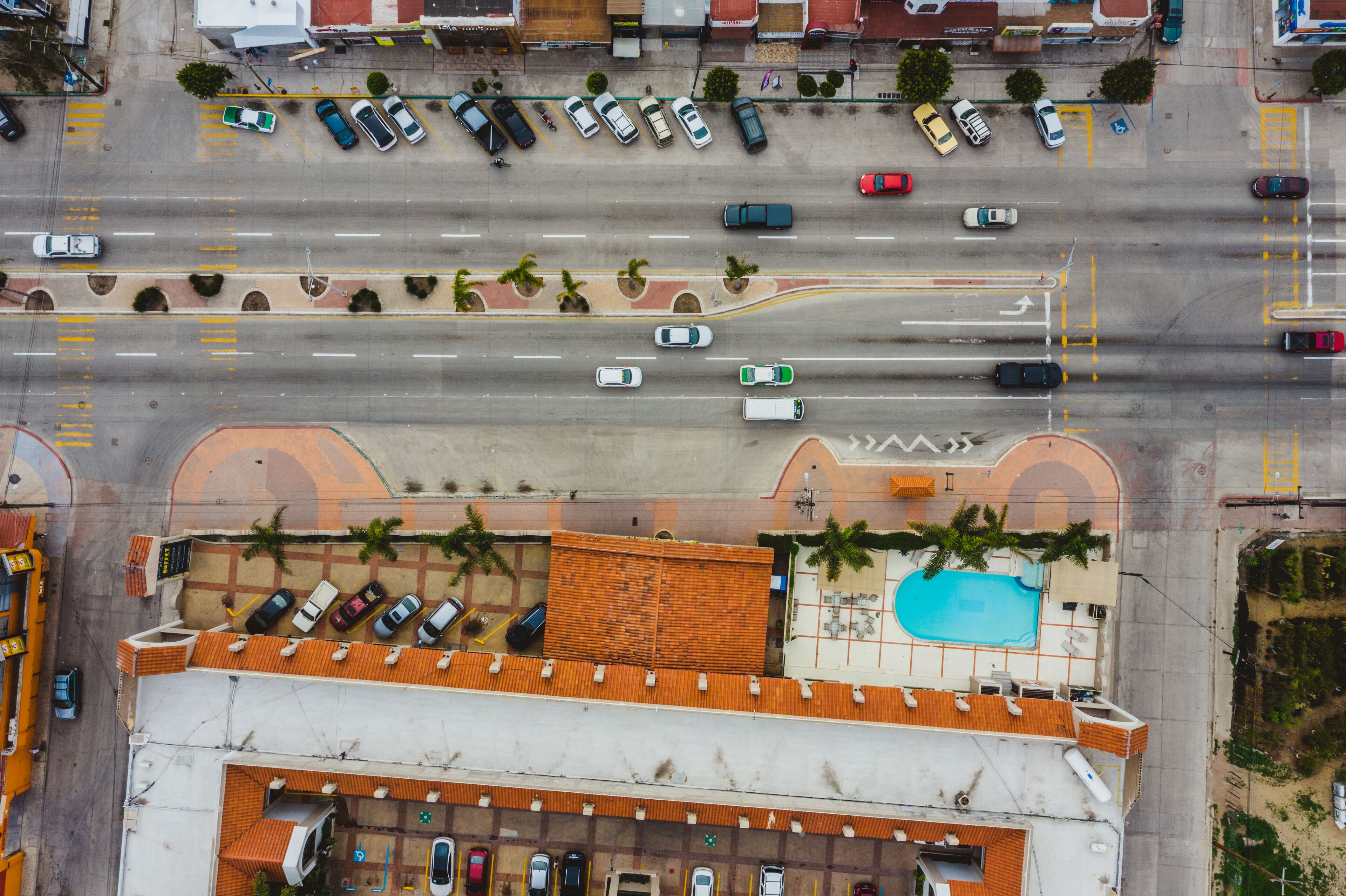 Vista aérea de uma rua com carros estacionados em ambos os lados, uma piscina e edifícios com telhados laranja.