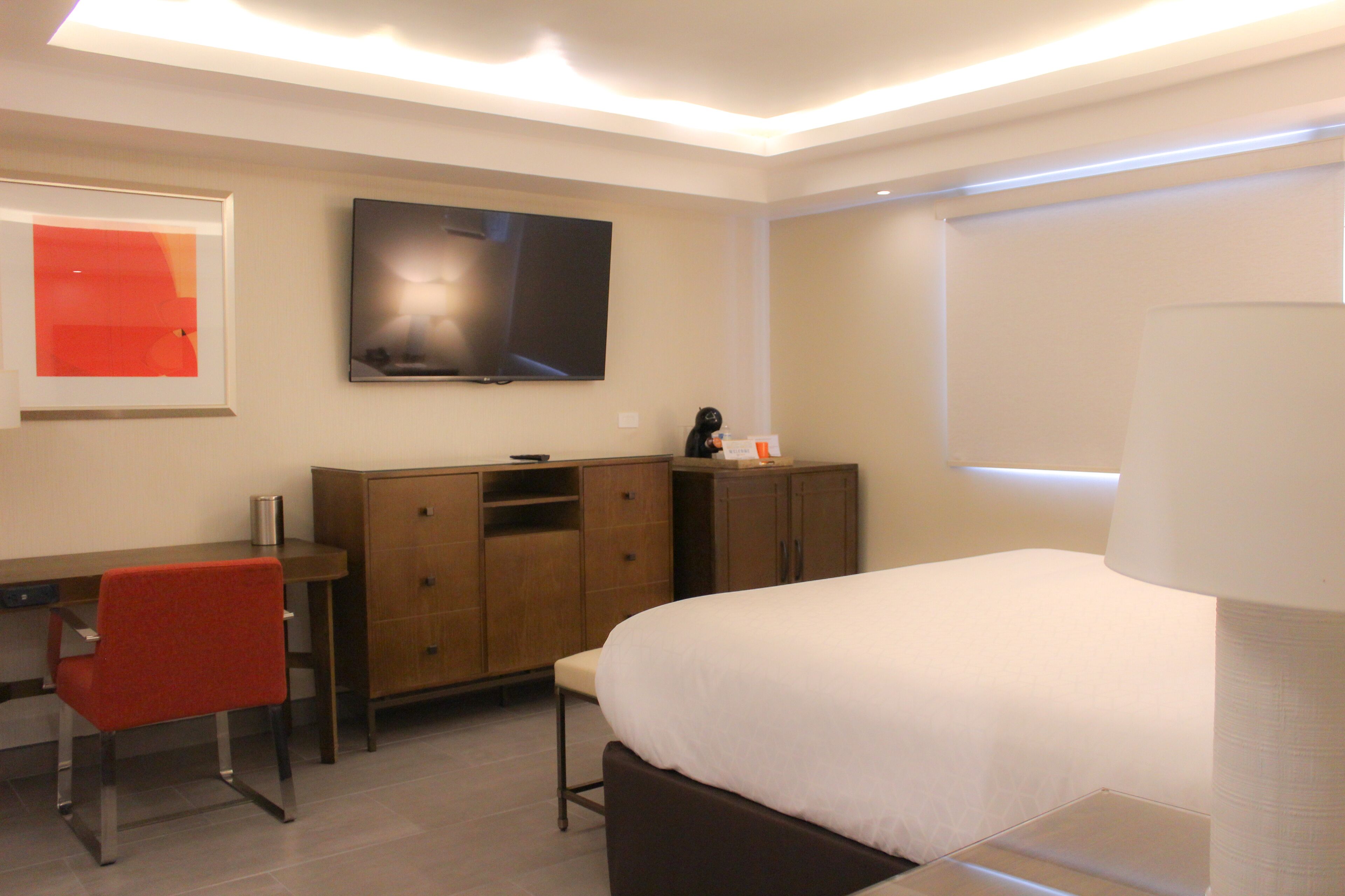 A modern hotel room with a bed, a desk, a chair, a television, and a cabinet.