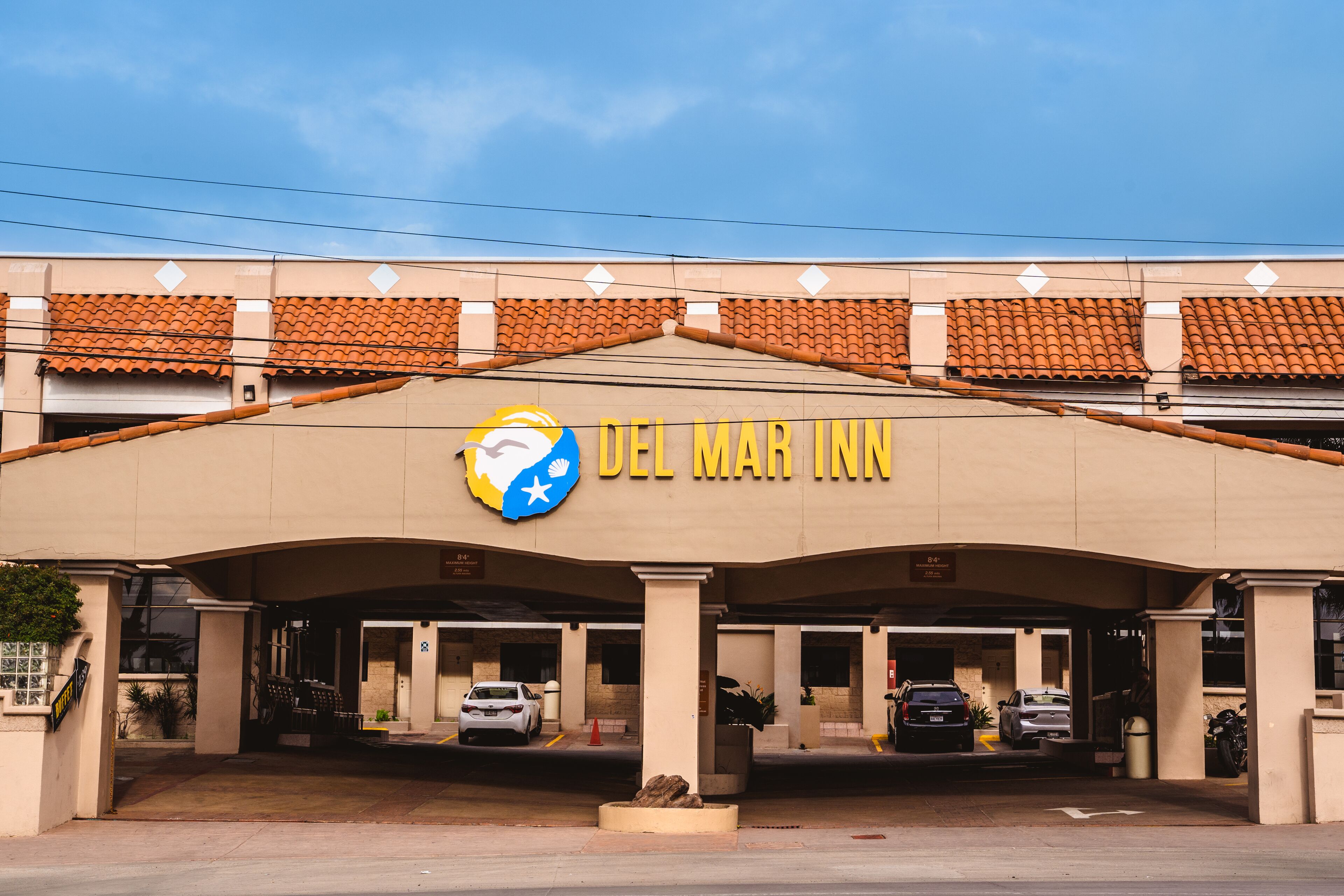 The exterior of Del Mar Inn with a tiled roof, a prominent logo, and a parking area with several cars.