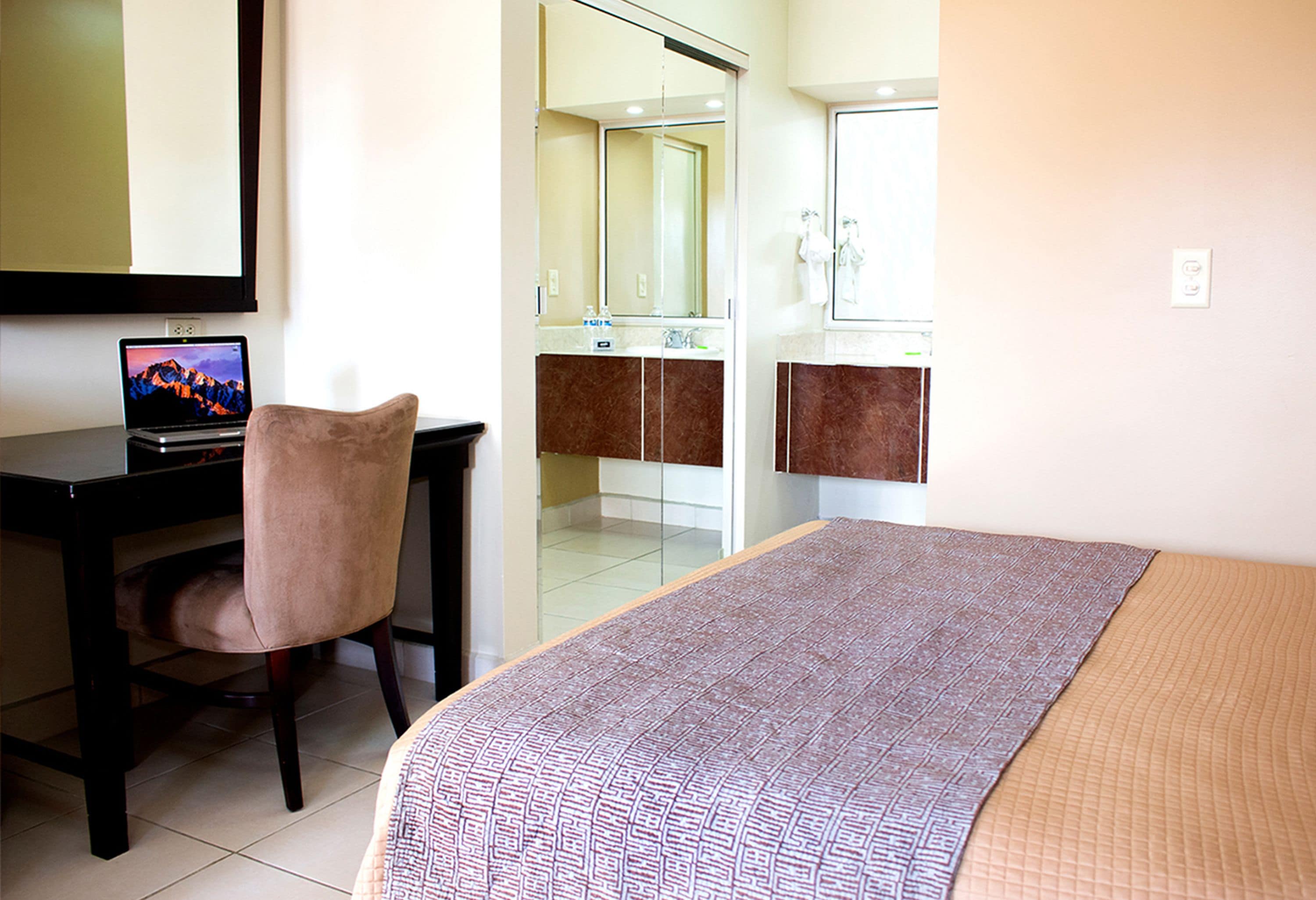 Habitación de hotel con cama, escritorio con computadora portátil, silla, baño con espejo y ducha.