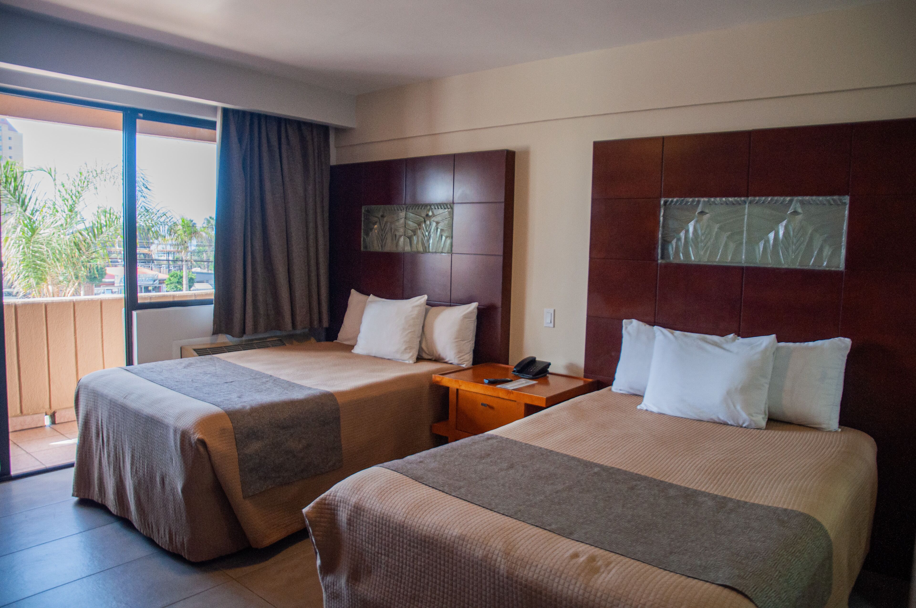Habitación de hotel con dos camas, cabecera de madera, mesita de noche y vistas al exterior.