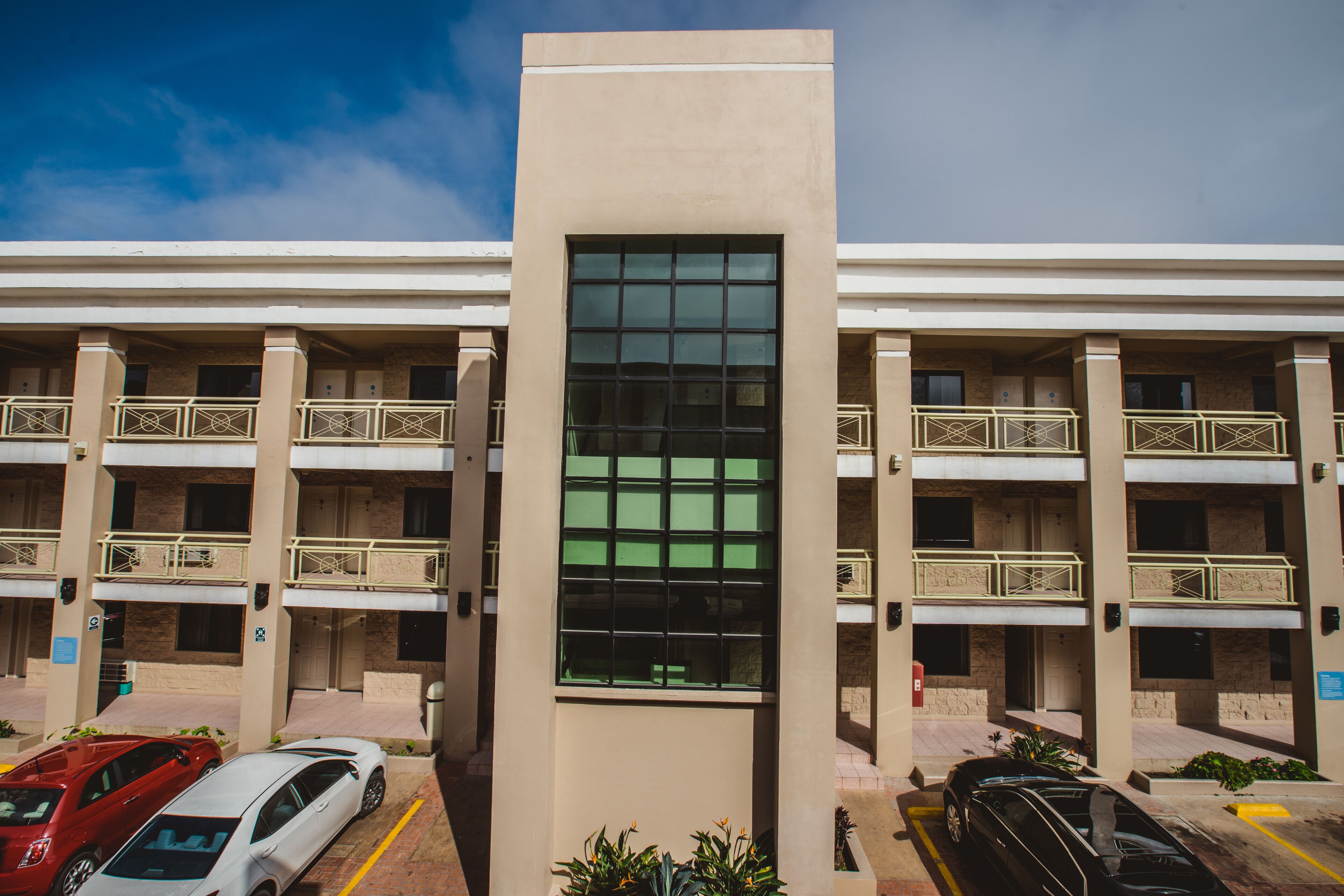 Un hotel de varios pisos con fachada de vidrio central, balcones y un estacionamiento con automóviles.
