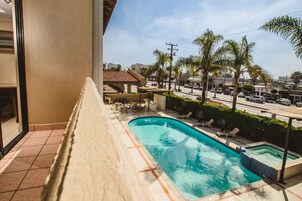 Una piscina con cubierta de baldosas, rodeada de sillones de descanso y palmeras, con vista a una calle y edificios al fondo.