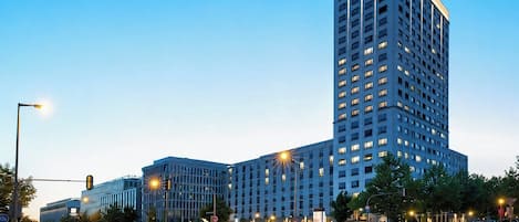 A multi-story hotel with a modern design, featuring a prominent corner building and a parking area with cars.