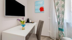 Une pièce avec un bureau blanc, une chaise, un téléviseur à écran plat fixé au mur, un petit vase avec des fleurs et un tableau représentant une fleur rouge, également accroché au mur.