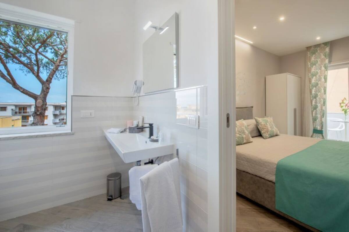 Ein modernes Hotelzimmer mit Bett, Badezimmer mit Waschbecken und Spiegel sowie ein Fenster mit Blick auf einen Baum.