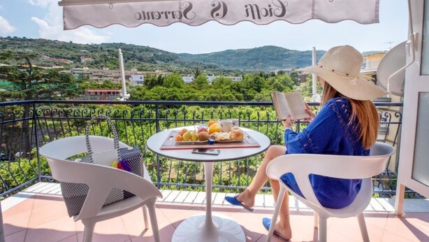 Une femme est assise sur un balcon, en train de lire, avec une table remplie de nourriture et une vue sur la campagne.