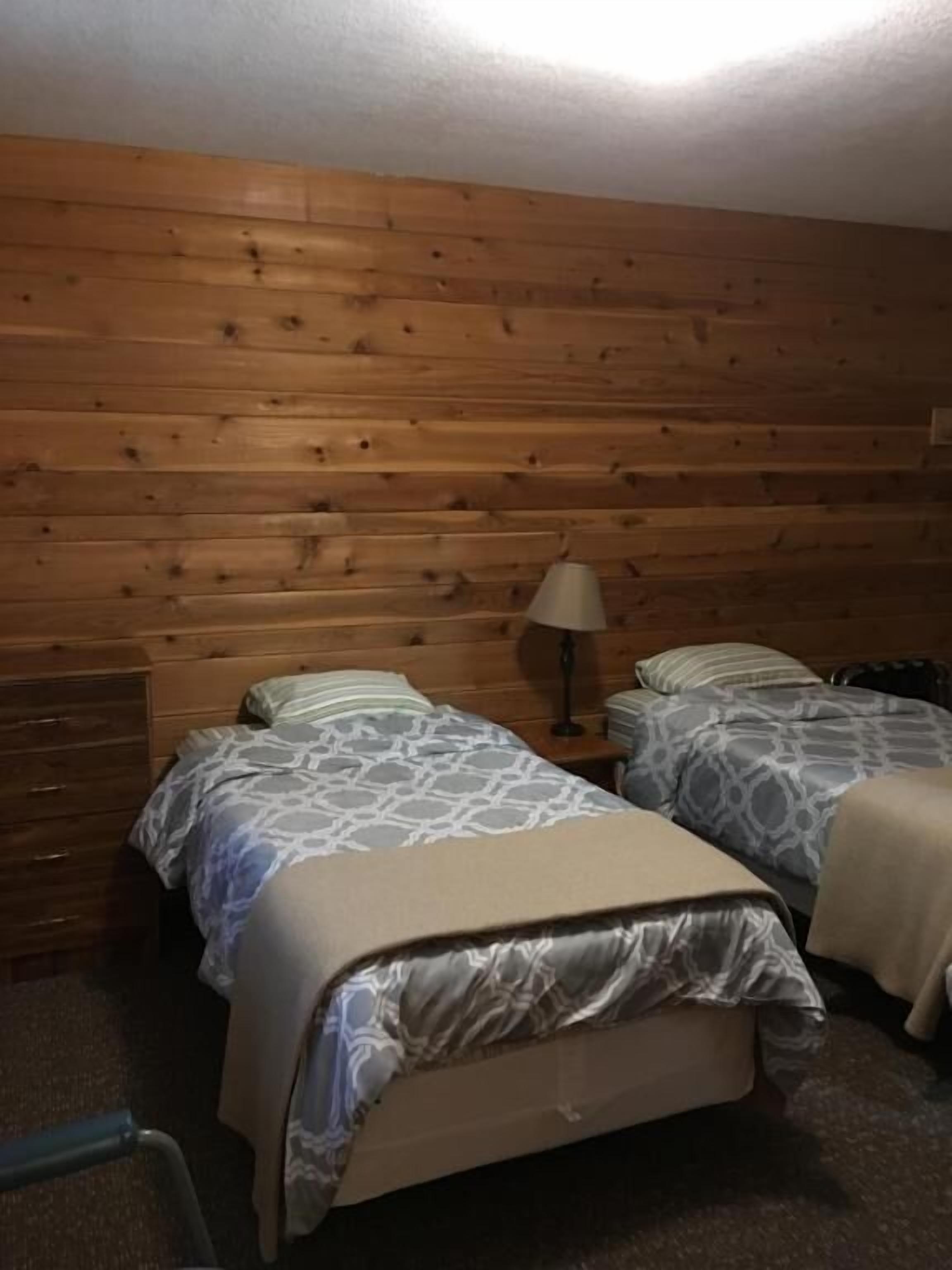 Ein Zimmer mit Holzwandverkleidung, zwei Betten mit gemusterten Laken, einem Nachttisch mit Lampe und einem Sessel.