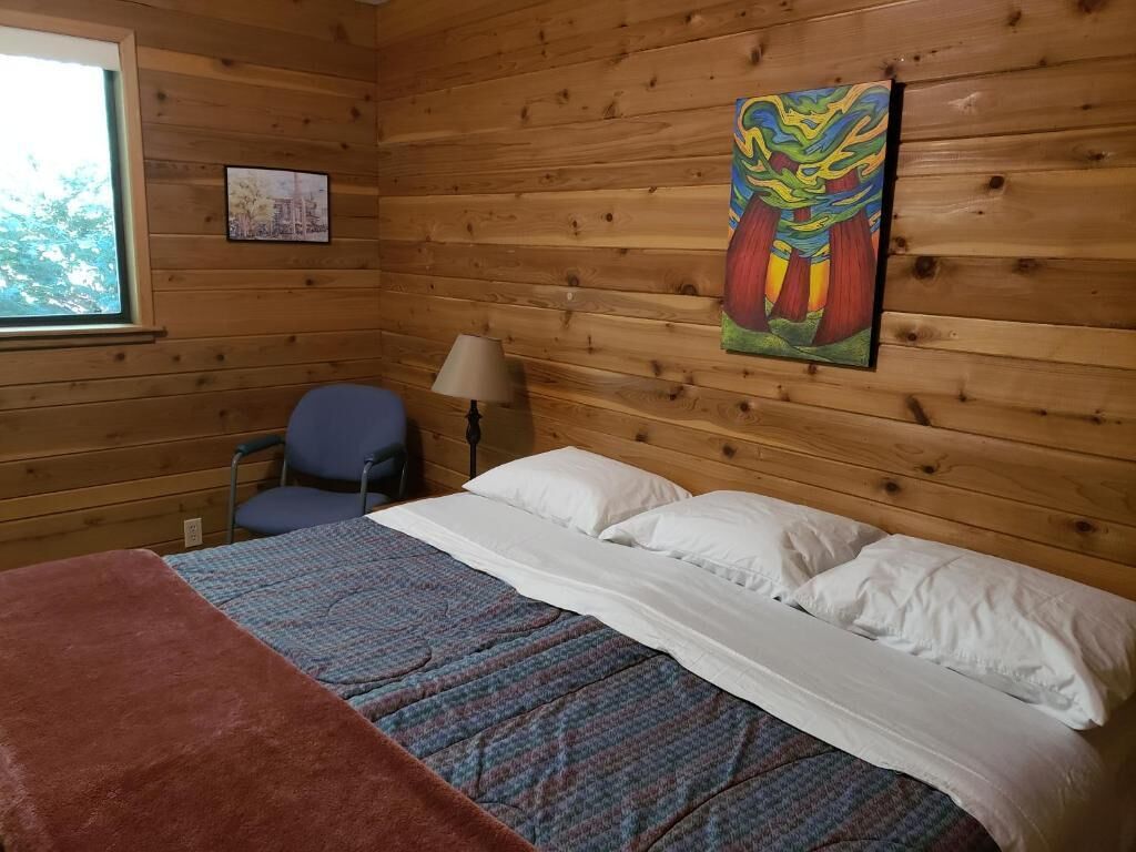 A wooden cabin room with a bed, a chair, a lamp, and a painting on the wall.