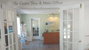 A reception area with a sign reading "The Centre Shop & Main Office," a reception desk, a fan, and a display of items on shelves. - Bethlehem Centre (Nanaimo)