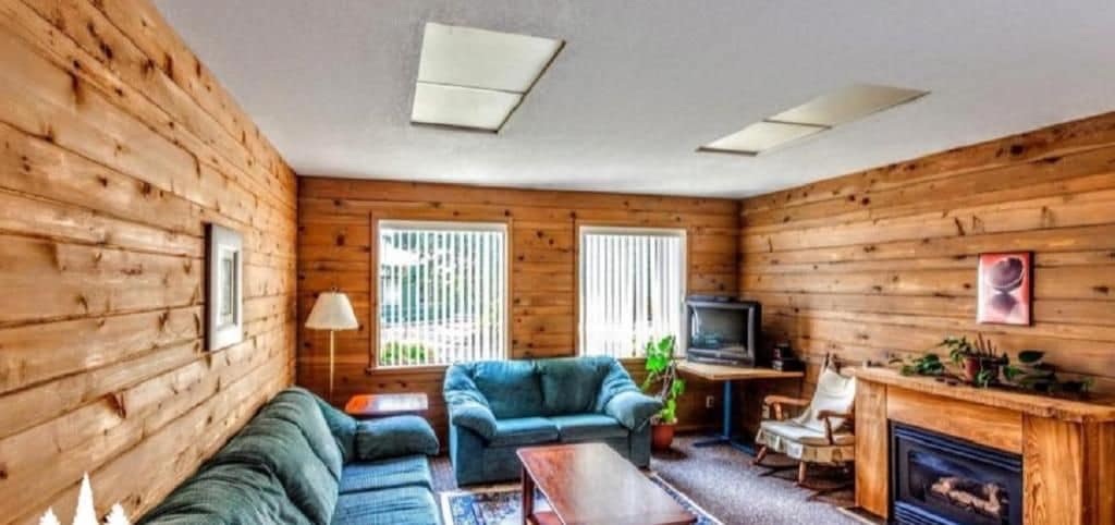 A cozy room with wooden walls, a sofa, a TV, a fireplace, and a window with blinds.