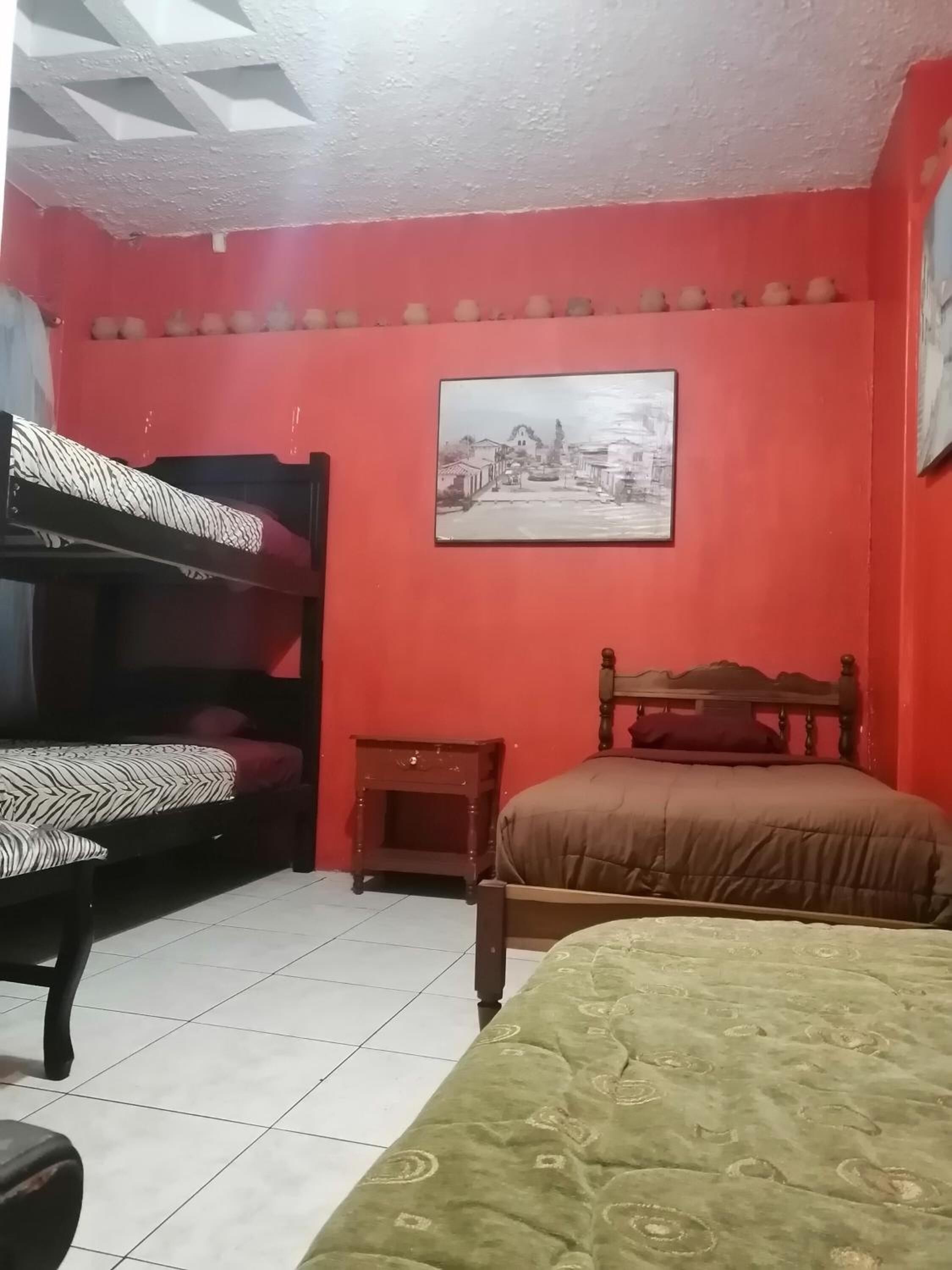 A room with a red wall, a bed with a brown headboard, a bunk bed with a zebra-patterned mattress, a small wooden nightstand, and a framed picture of a cityscape.