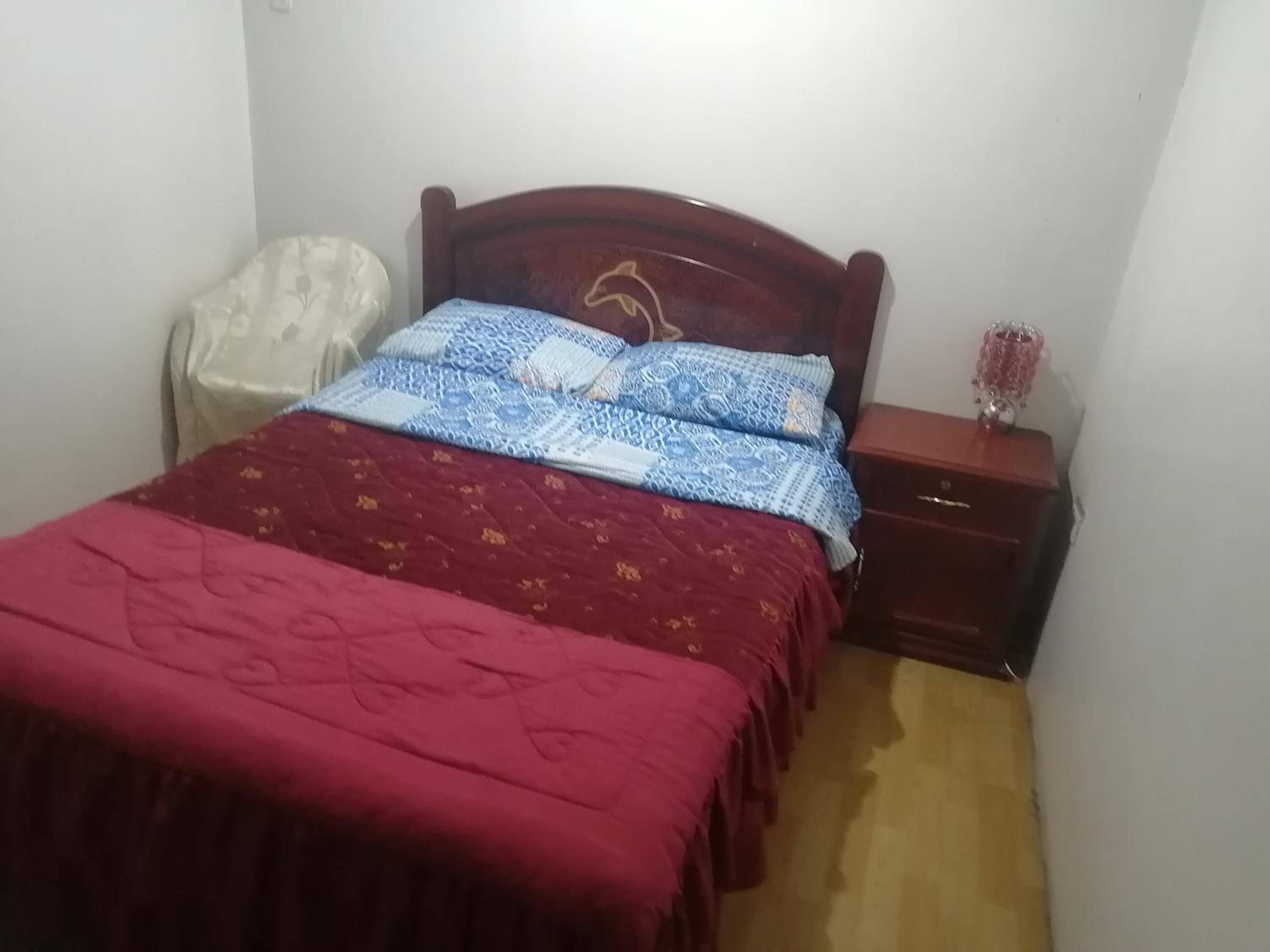 A bedroom with a wooden bed, a red bedspread, a blue and white patterned bedspread, a white chair, and a nightstand with a pink vase.