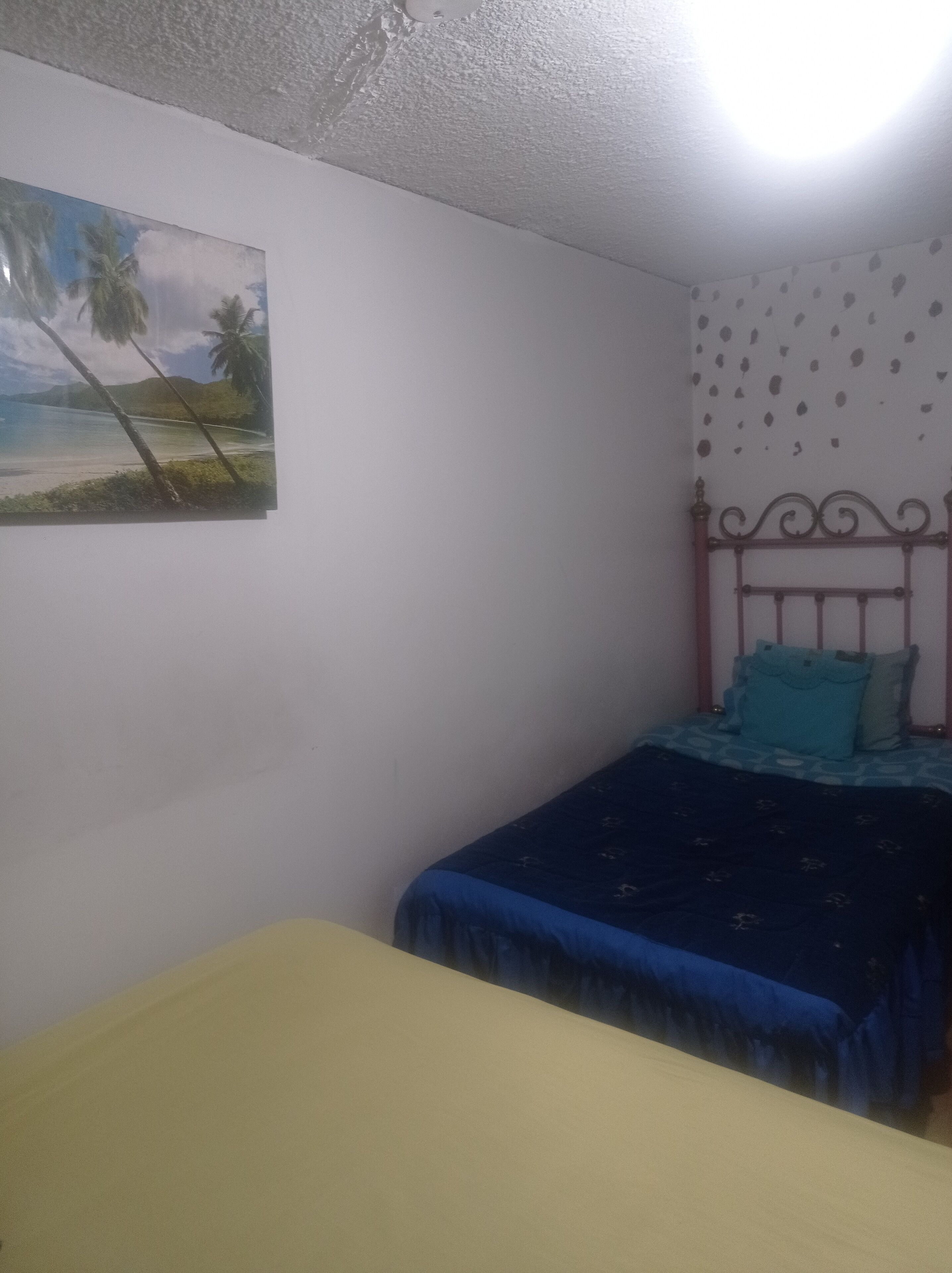 Un dormitorio con una cama, un sofá amarillo, una pared con un cuadro tropical y una lámpara de techo.
