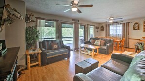 A spacious living room with a large sofa, a smaller sofa, a coffee table, and a dining area with a table and chairs. There are two ceiling fans, a large window with curtains, and a plant. - Waterfront Home w/ Fire Pit on Table Rock Lake (Golden)