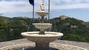 Una fuente de piedra de varios niveles y con banderas, rodeada por un área empedrada con vistas a una colina y edificios al fondo.
