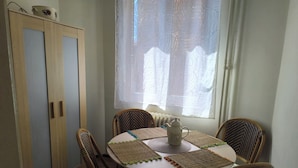 Un'area pranzo con un tavolo rotondo, quattro sedie in vimini, una tovaglia bianca, un teiera bianca e una finestra con tende leggere.