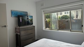 Ein Schlafzimmer mit einem Bett, einem Schminktisch, einem Fernseher an der Wand und einem Fenster mit Blick nach draußen.