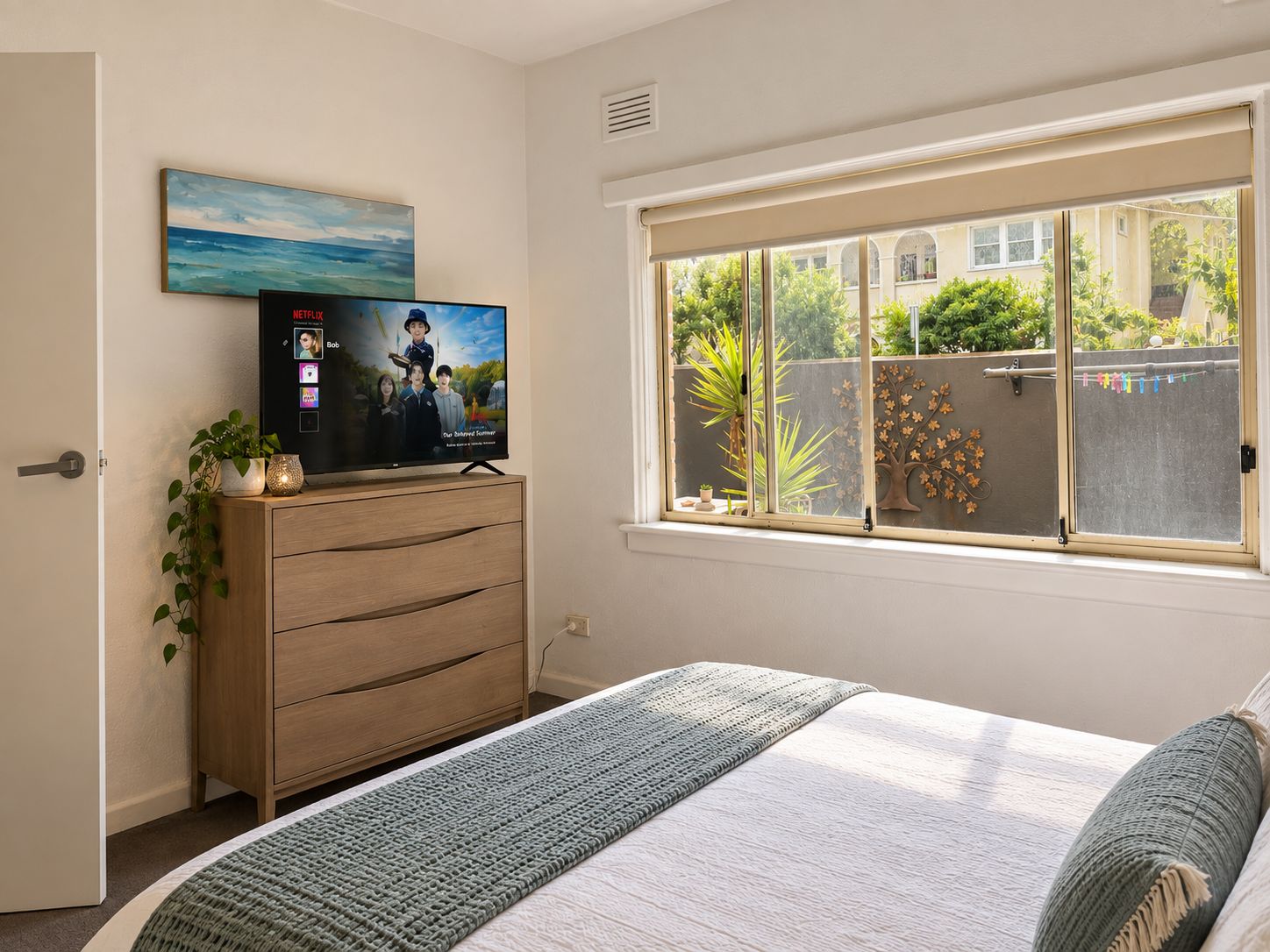 Une chambre à coucher avec un lit, un téléviseur posé sur une commode en bois, une fenêtre donnant sur un paysage verdoyant et un tableau accroché au mur.