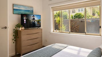 Une chambre à coucher avec un lit, un téléviseur posé sur une commode en bois, une fenêtre donnant sur un paysage verdoyant et un tableau accroché au mur.