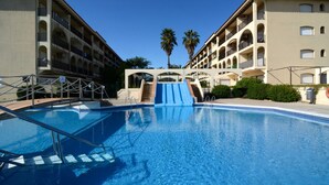 A swimming pool with a blue slide, surrounded by a multi-story building with balconies and palm trees. - JARDINS DEL MAR 114 (Estartit)
