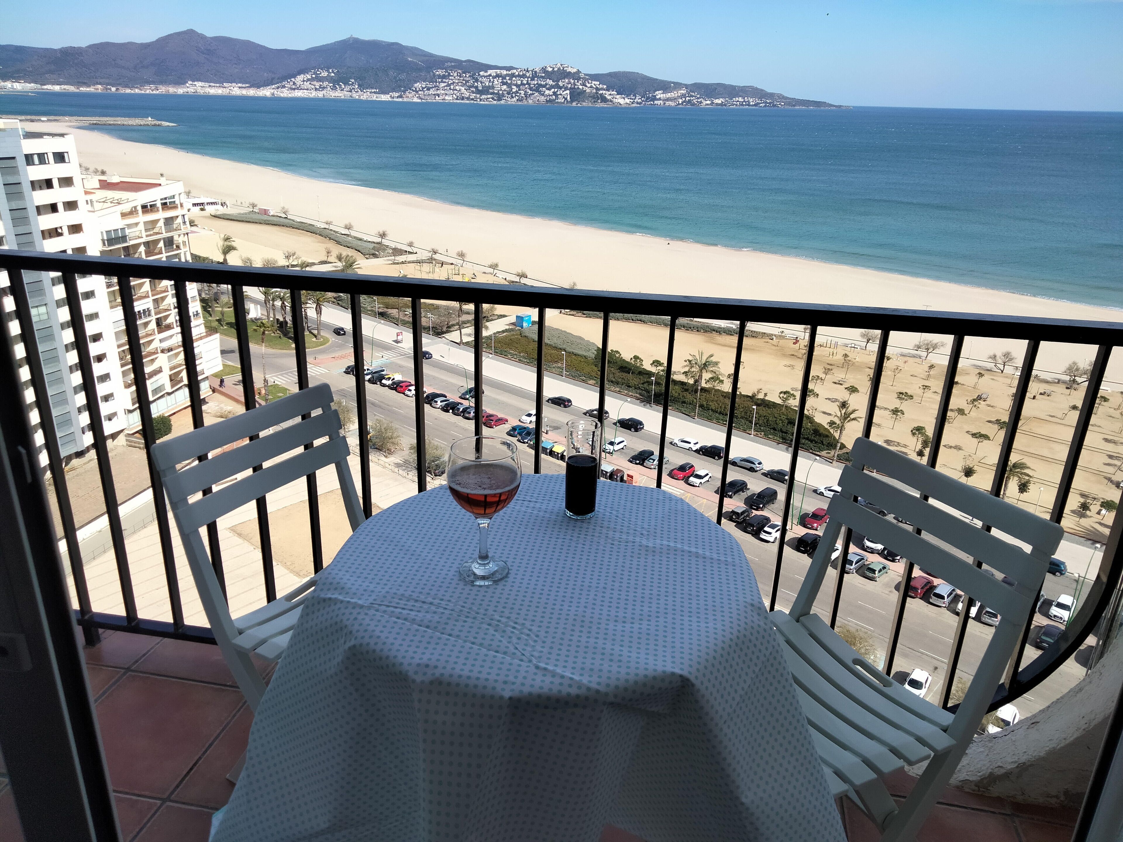 A balcony with a table and chairs overlooking a beach and a parking lot.