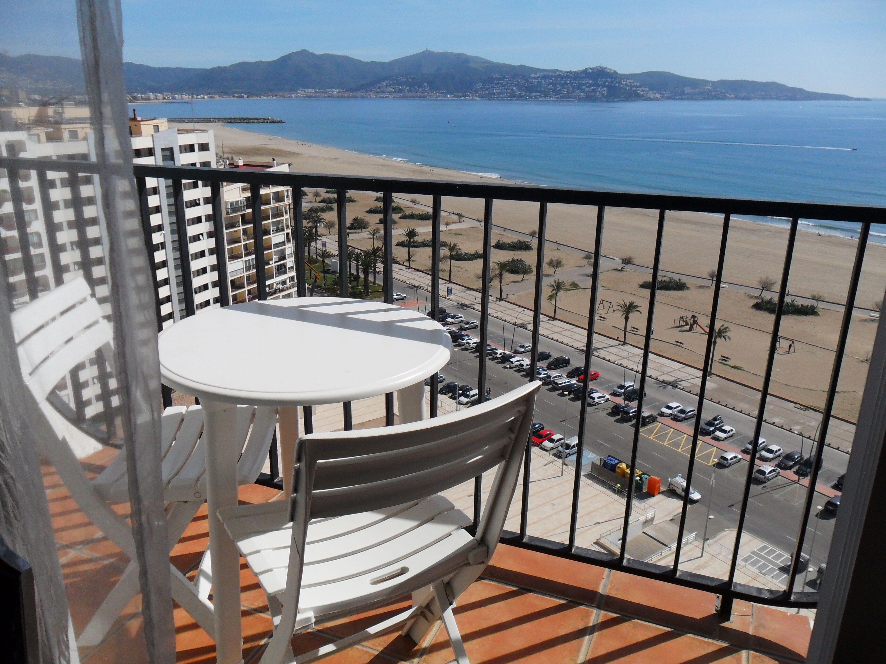 Varanda com mesa e cadeiras, vista para uma praia e um estacionamento.