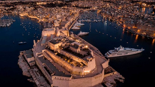 An aerial view of a historic fortified city with a prominent castle-like structure surrounded by water, boats, and a marina.