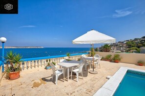 A terrace with a pool, white tables and chairs, a white umbrella, and a view of the sea. - Villa Ocean in Mellieha with Fantastic Sea Views 4 bedrooms A/C Sleep 9 people  (St Maria Estate)