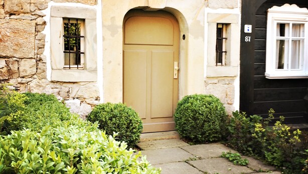Una casa tradicional con puerta color beige, paredes de piedra y un pequeño jardín con arbustos.
