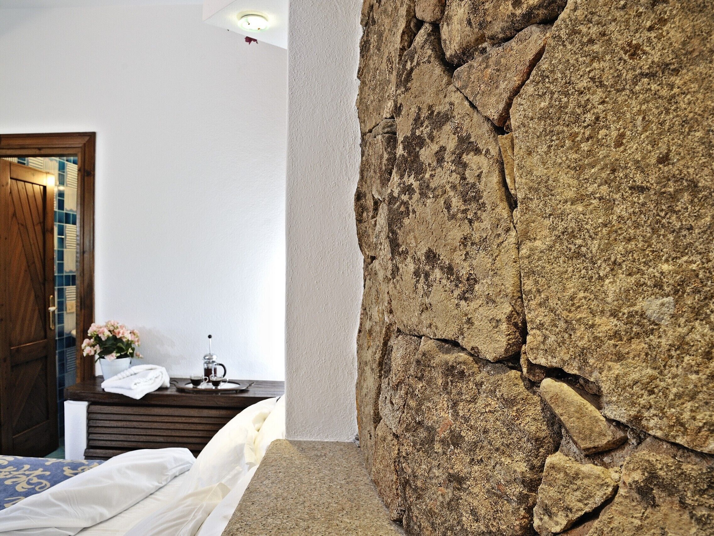 Un dormitorio con pared de piedra, puerta de madera y una mesita de noche con un florero.