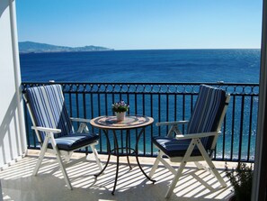 Un balcon avec deux chaises à rayures, une petite table ronde ornée d’un pot de fleurs, et une vue sur la mer.