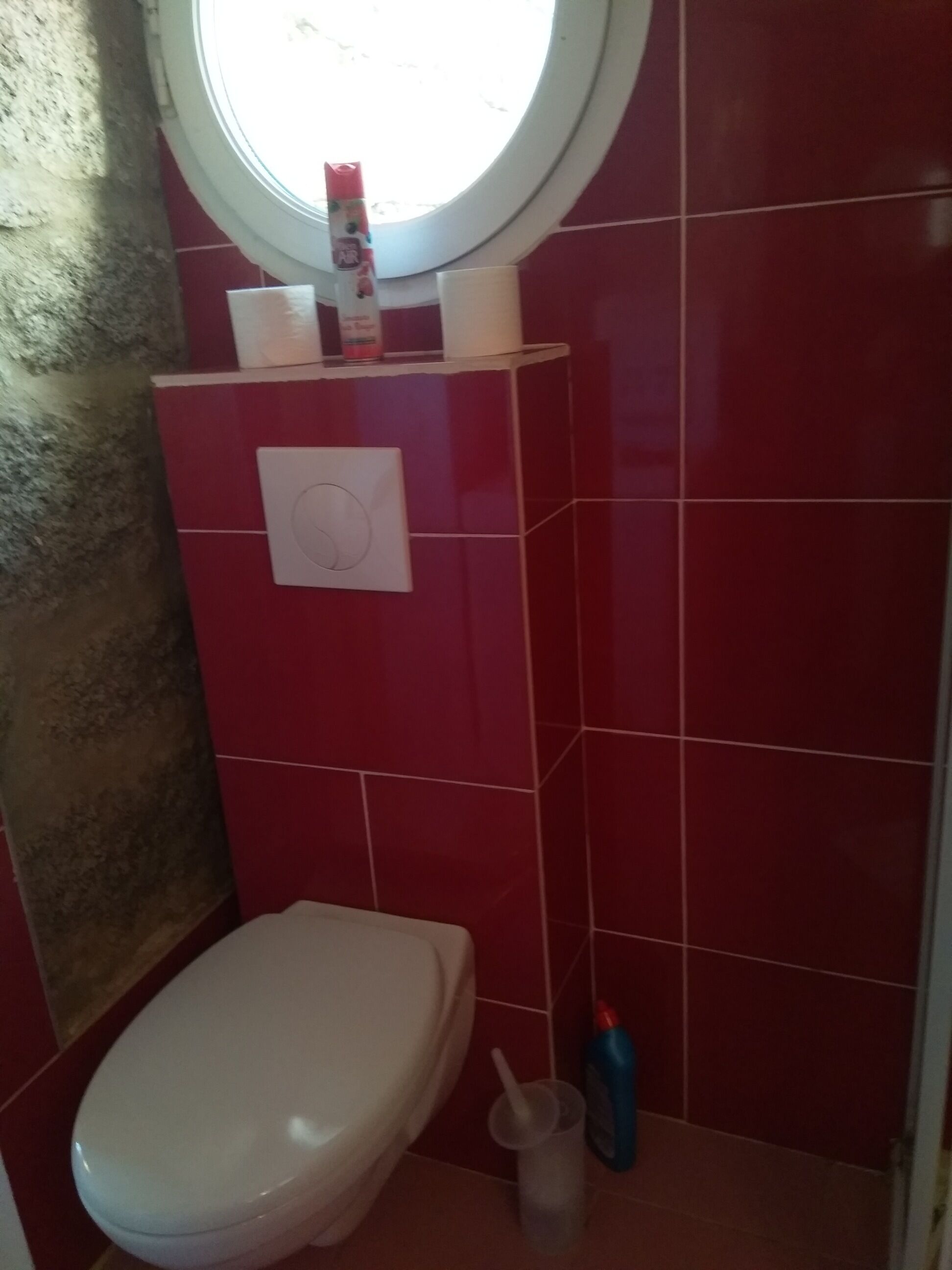 Une salle de bain avec des toilettes blanches, une fenêtre ronde, des murs recouverts de carreaux rouges, une étagère blanche contenant des articles de toilette et un porte-rouleau de papier toilette blanc.