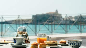 Une table dressée pour le petit-déjeuner, avec des viennoiseries, une tasse et un verre de jus d’orange, placée au bord de l’eau, offrant une vue sur un pont et des bâtiments.
