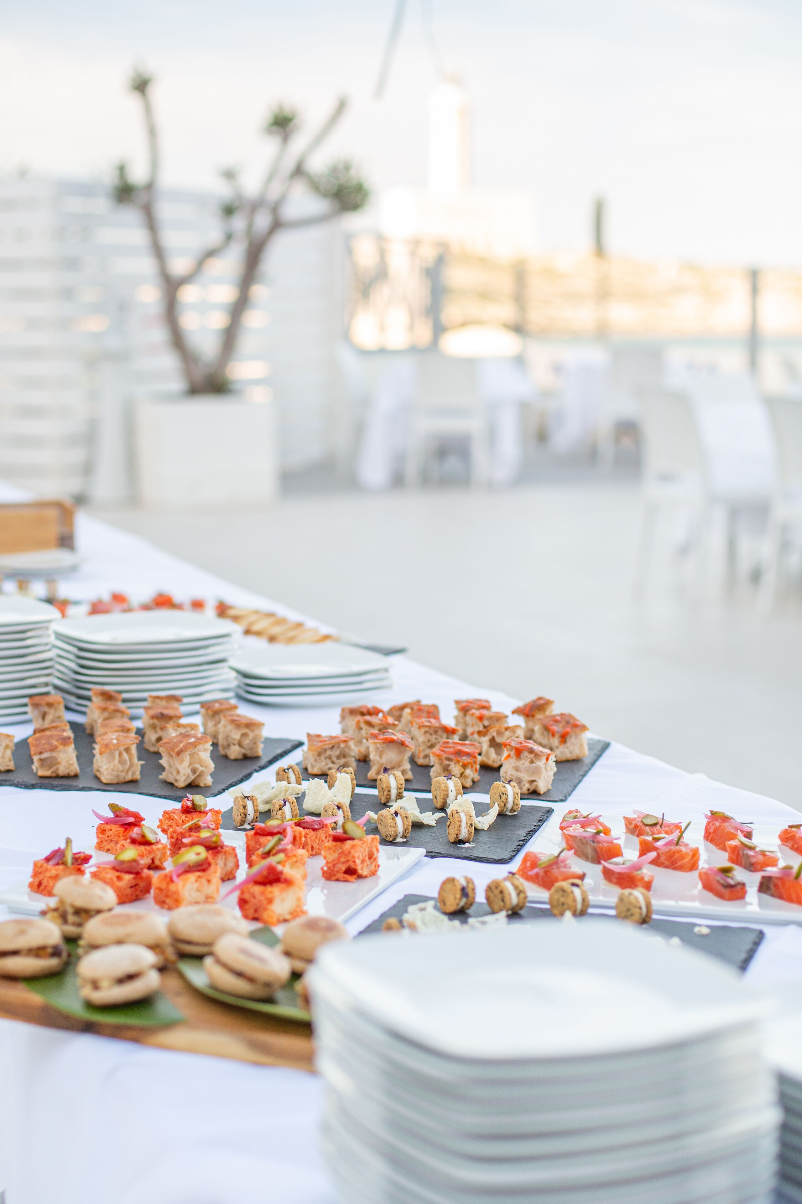 Uma mesa de buffet com diversos alimentos e pratos brancos empilhados.