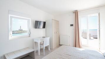 Een slaapkamer met een bed, bureau, stoel, televisie, kast, radiator en een balkon met gordijnen.